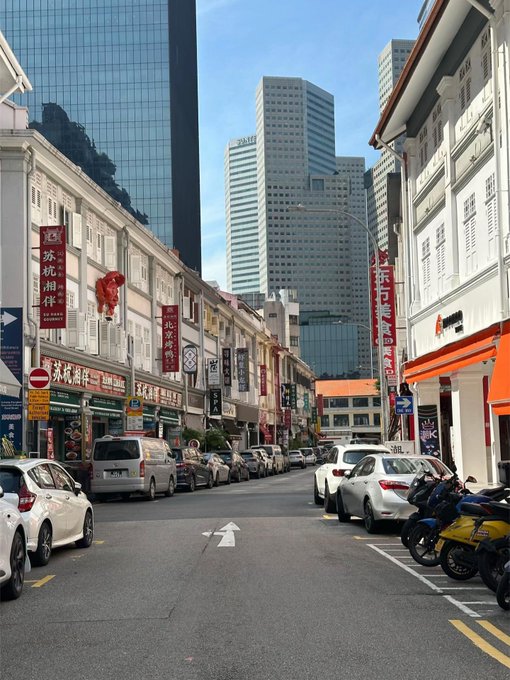 A street scene in Singapore's Chinatown with traditional shophouses, parked cars, and modern skyscrapers in the background, featuring signs in Chinese including 沫城商伴 (Mocheng) and 上海银行 (Bank of Shanghai).