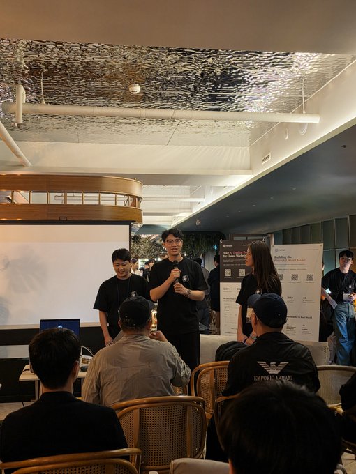 A man in a black t-shirt speaks into a microphone while presenting at an indoor event, with a research poster visible in the background and an audience seated on rattan chairs.