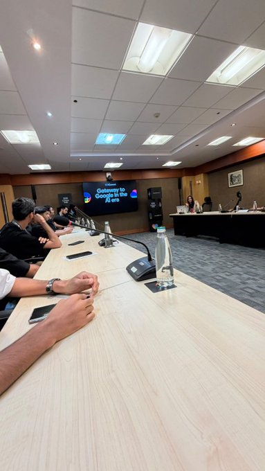 A conference room setting with people seated at a long table, with a screen displaying the Google logo and text 'Gateway to Google in the AI era'.