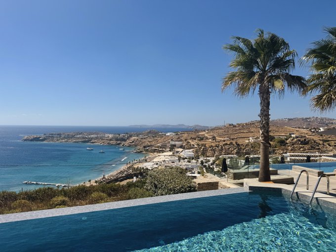 A stunning coastal view from a villa pool area features blue water, a palm tree, and a distant coastline under a clear sky.