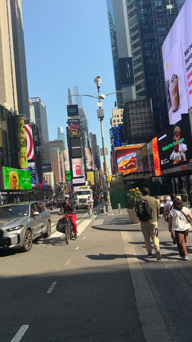 A street scene in Times Square, New York, featuring tall buildings covered in digital billboards, pedestrians walking, and a cyclist in red pants riding alongside a car.