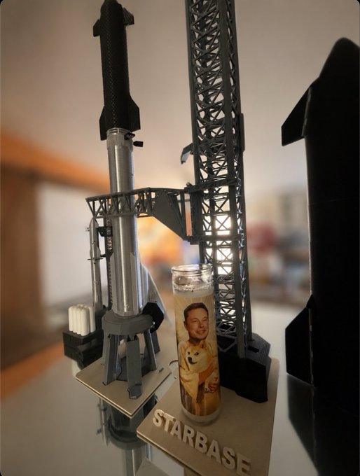 A model of the SpaceX Starship rocket on a "STARBASE" launch pad display, with a prayer candle featuring Elon Musk holding a Shiba Inu (Doge) placed in front, humorously symbolizing the "Doge to the Moon!" mission to send the meme-inspired cryptocurrency to lunar heights aboard Musk's rocket.