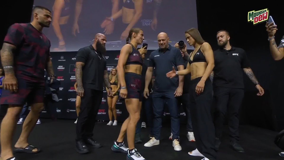 Two female UFC fighters, Marina Spasic and Stephanie Luciano, stand face-to-face in a tense staredown at the UFC Belgrade weigh-in event, flanked by security personnel and officials including Dana White in the center. The "Mountain Dew" sponsor logo is visible in the upper right corner, with UFC branding throughout the backdrop.
