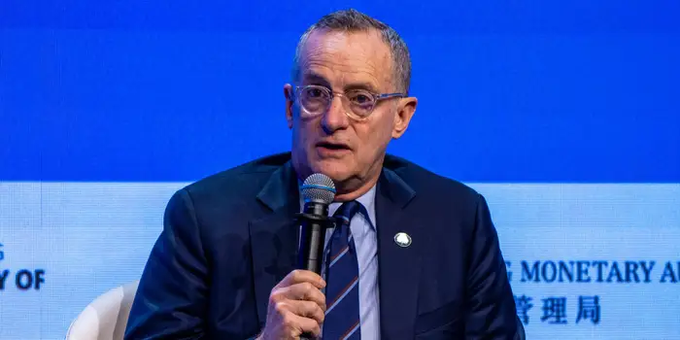 An older man with glasses and a dark suit speaks into a microphone at a conference, with a blue screen behind him displaying partial text 'MONETARY A' and Chinese characters '管理局'.