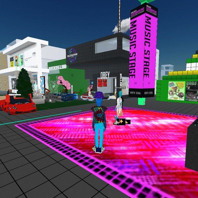 An avatar stands before a glowing pink stage in a vibrant blockchain-themed virtual world, featuring prominent "MUSIC STAGE" signage and a building labeled "OBEY," capturing the lively atmosphere of the reported Voxels event around the Blockchain Radio and "$traylight" locations.