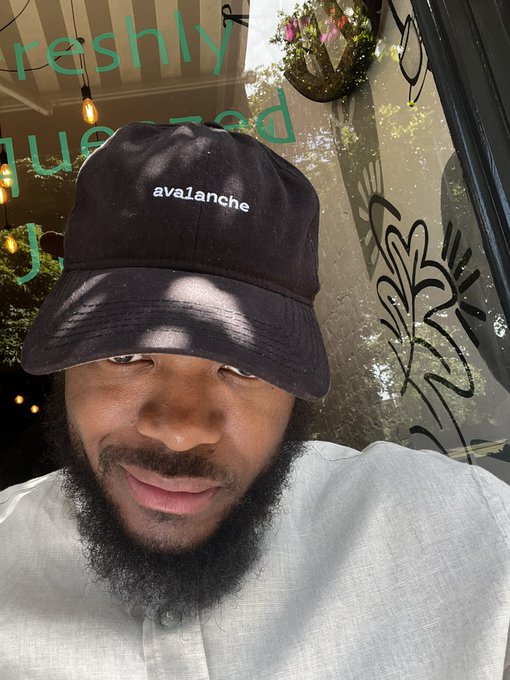 A man wearing a black bucket hat with "avalanche" embroidered in white smiles in a selfie taken outside a café with window text reading "freshly squeezed." The image ties to the post by visually representing the Avalanche ecosystem affiliation referenced in the announcement about the Collaboration Cohort and Builder Grants.