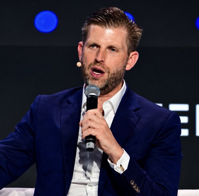 Eric Trump, wearing a blue suit and white shirt, speaks into a microphone while seated on a panel.