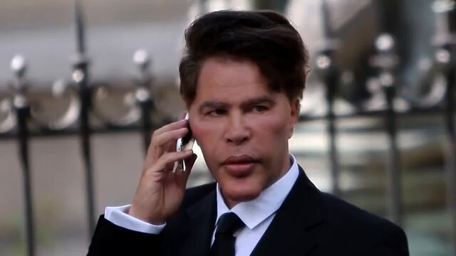 The image features a man dressed in a suit, talking on his cell phone while standing outside of a building with a gate or fence behind him. The man appears to be engaged in a conversation, possibly discussing the recent news about Ripple XRP holders not listening to reality checks and holding onto their digital assets.
