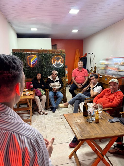 A group of people sitting in what appears to be a small restaurant or café, with one man in the foreground gesturing while others watch, near a green wall with neon signs.