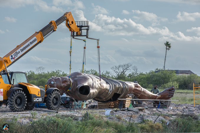 A construction crane lifting a large bronze statue of a reclining figure at an outdoor site with rubble and a palm tree in the background.