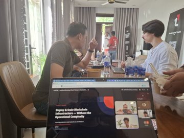 Two people are having a conversation at a table with water bottles, while a laptop in the foreground displays a presentation slide titled "Zephyr & Anka Blockchain Infrastructure — Without the Operational Complexity" by ZKAS.
