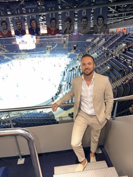 A man in a cream-colored suit stands smiling on the steps of an indoor arena overlooking an empty ice hockey rink with championship banners hanging above.