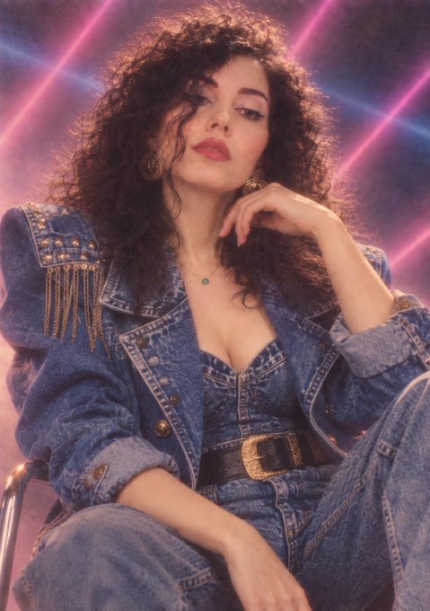 Fashion editorial portrait of a woman with voluminous curly hair wearing a denim outfit with shoulder fringe, posed against a neon pink and blue background.