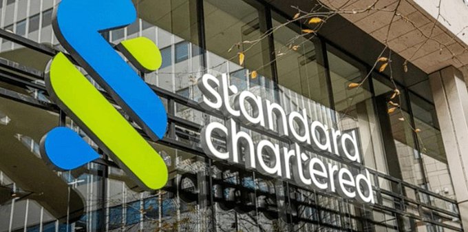 The exterior signage of Standard Chartered bank, featuring their blue and green interlocking logo next to the company name in white lettering on a glass building facade.