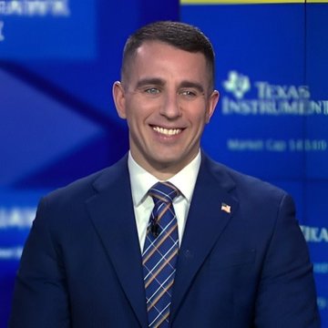 A man in a dark blue suit, white shirt, and striped tie smiling, standing in front of a Texas Instruments backdrop during what appears to be a debate or broadcast.
