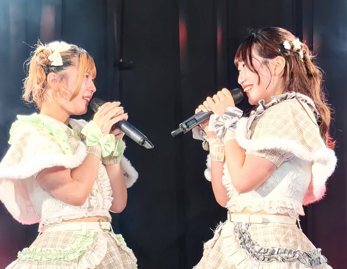 Two young women in matching frilly pastel idol costumes with bow hair accessories smile while facing each other on stage, each holding a microphone.