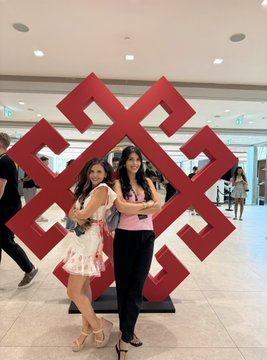 Two women pose together in front of a large red geometric sculpture featuring a stylized cross-like design at the Solana Summit Serbia. The setting appears to be a modern indoor venue with polished floors, suggesting a conference or event space.