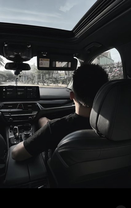 A view from inside a car showing a person with short dark hair in the driver's seat, with a sunroof open and a street scene visible through the windshield.