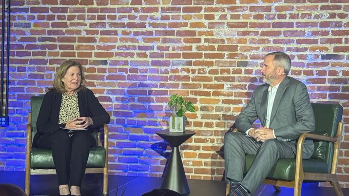 A woman and Brad Garlinghouse are seated in chairs on a stage with an exposed brick wall background, engaged in what appears to be a fireside chat or interview discussion, likely addressing topics related to the CLARITY Act and crypto regulation.