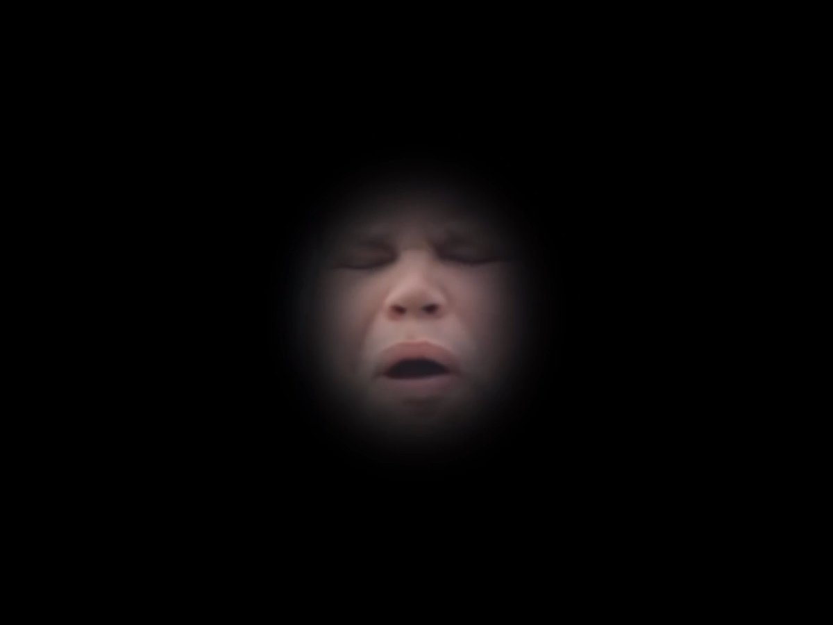 A child's face emerges from a dark background through a circular peephole, eyes closed and mouth slightly open as if blowing a kiss.