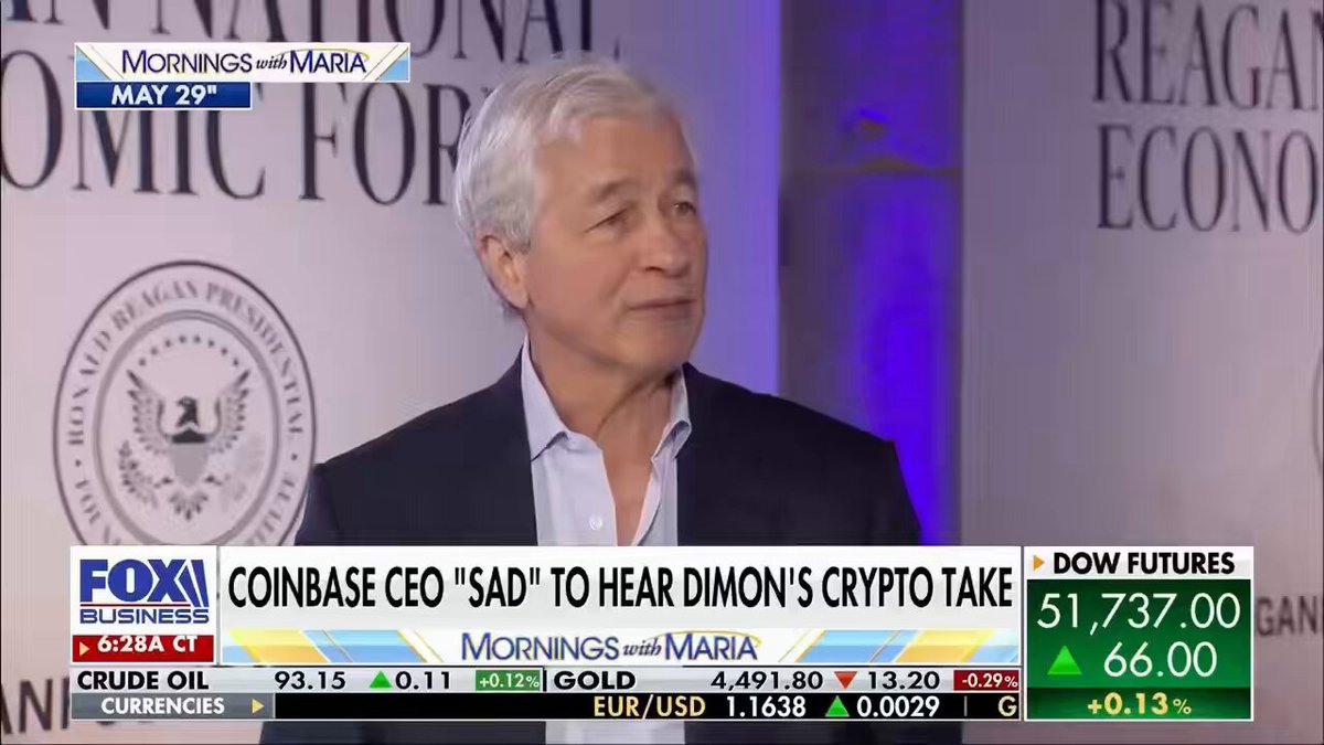 The image shows a man wearing a suit standing in front of a TV screen, with a news anchor reporting on a conversation between two men about cryptocurrency. The news anchor is discussing the CEO of Coinbase, Brian Armstrong, who is being criticized by Jamie Dimon for his views on Bitcoin and crypto. The post also mentions that the CEO is sad to hear Dimon's take on the matter.