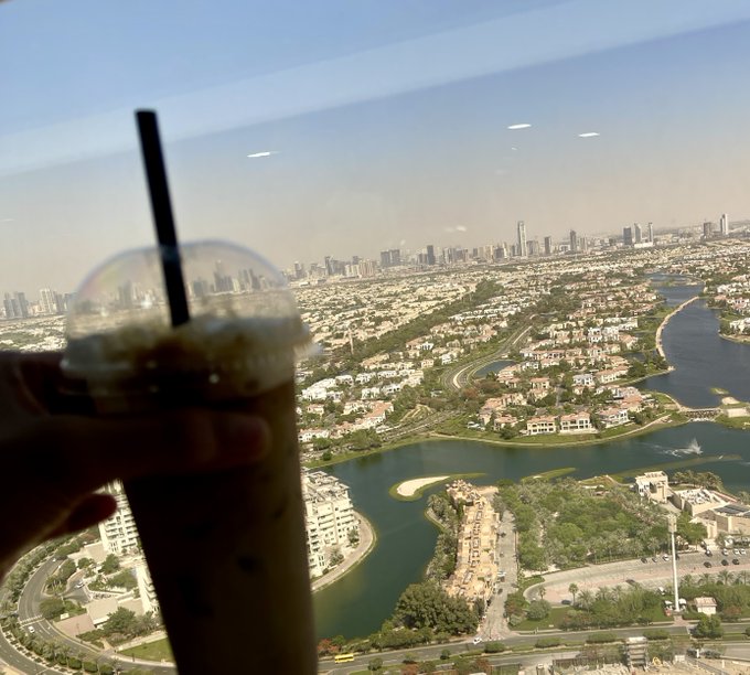 A hand holding an iced coffee or tea with a black straw in the foreground, with a panoramic aerial view of a city skyline, lake, and residential areas visible through a window.