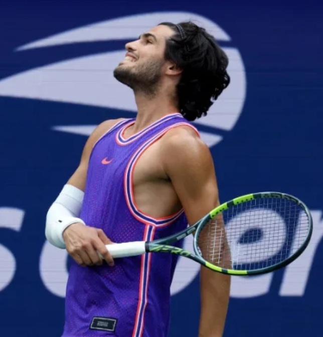 A tennis player in a purple Nike tank top with red and white trim holds a racket and looks upwards, appearing emotional, with a 'SO' logo visible in the blue background.