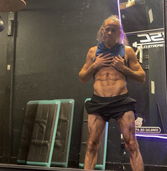 A very lean, muscular older man with long gray hair poses in a gym mirror, pulling open his blue sleeveless hoodie to reveal his abs, with reversed text visible on the wall.