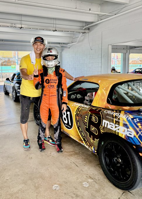 A man in a yellow shirt posing with a young child in an orange racing suit and helmet next to a yellow Mazda race car (#81).