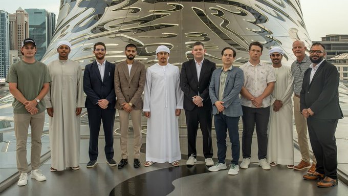 The image shows a group of men posing for a photo in front of an outdoor sculpture or landmark, possibly the Keeta joint venture between Saudi Arabia and Israel as described in the caption provided. The photograph emphasizes unity, collaboration, and shared goals among the members present. Each man in the group is dressed formally, wearing suits and ties, adding to the overall sense of professionalism and seriousness associated with theKeeta project.