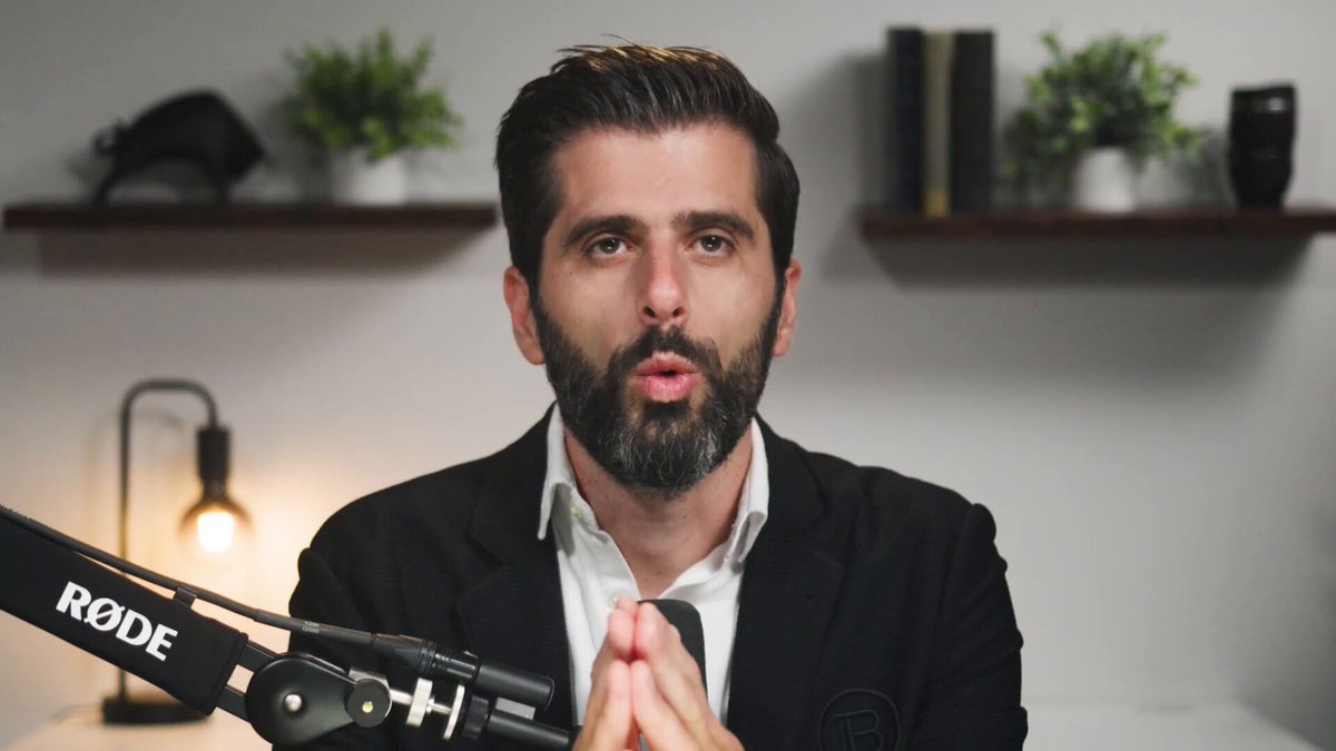 The image features a man wearing a suit, sitting at a desk with a microphone in front of him. He has a beard and mustache, giving him a distinguished appearance. The background includes several potted plants placed around the room, adding a touch of greenery to the scene. There is also an open laptop on the desk next to the microphone, suggesting that he might be involved in some form of online communication or presentation related to cryptocurrency.
