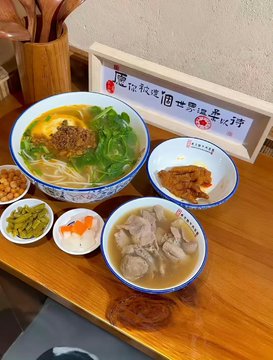 A top-down view of a Taiwanese meal featuring a bowl of beef noodle soup, a side of meat, pickled vegetables, and rice, with a small sign containing Chinese characters.
