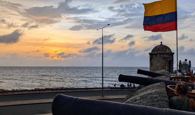 A scenic sunset view over the ocean featuring a Colombian flag flying alongside historical cannons and a stone sentry box.