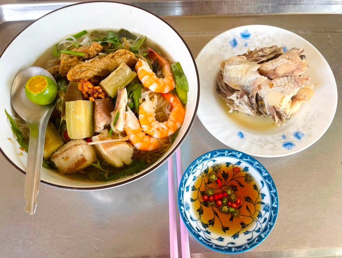 A Vietnamese meal featuring a large bowl of noodle soup with shrimp, meat, and herbs with a calamansi half, alongside a plate of boiled chicken and a small bowl of fish sauce with sliced chilies.