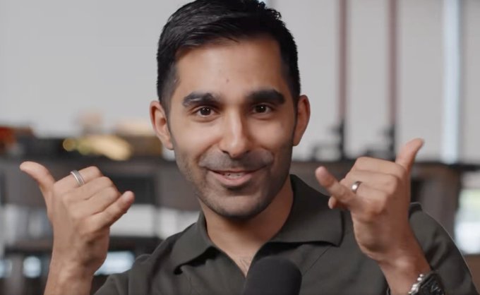 A close-up of a smiling man with dark hair and a short beard, wearing a dark shirt, who points both thumbs toward himself while wearing rings and a watch.