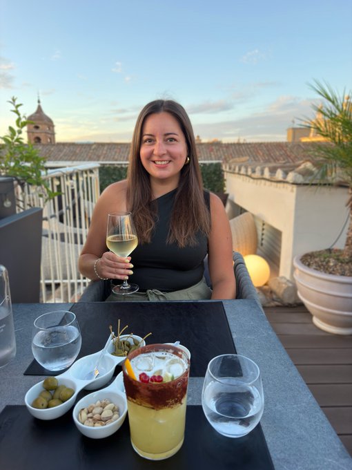 A young woman smiling on a rooftop terrace at sunset, holding a glass of white wine, with cocktails, olives, and mixed nuts on the table in front of her.