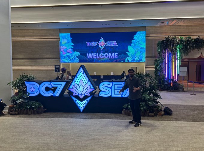 A man stands in front of a 'DG7 SEA WELCOME' sign at an event venue, with decorative plants and neon lighting.
