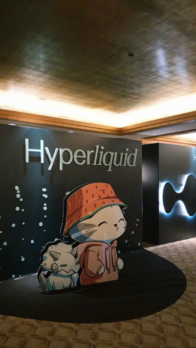 An indoor display for the 'Hyperliquid' brand features cartoon character mascots with an orange basket-like hat on a black wall.