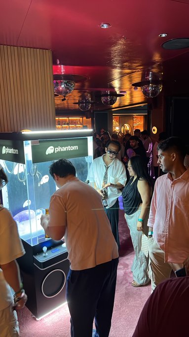 A crowd gathers around a 'Phantom' branded arcade-style machine at what appears to be an indoor event or venue with disco balls and pink lighting.