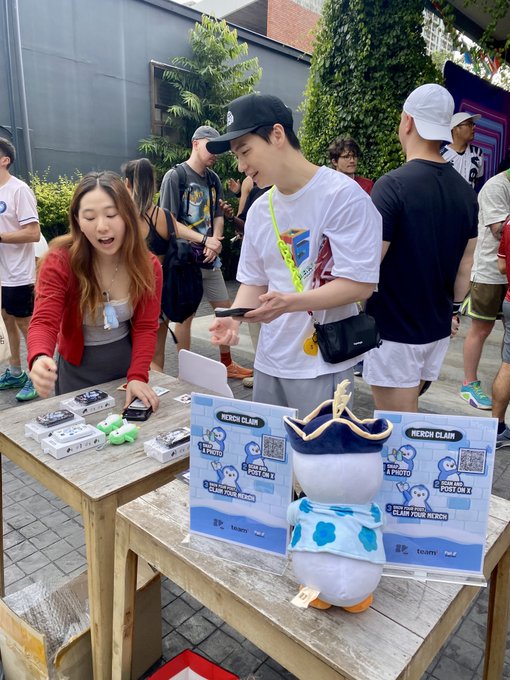 An outdoor merchandise booth displaying merchandise claim signs and plush toys, with two people interacting at the table.