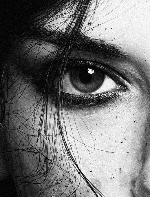 A dramatic black and white close-up photograph of a person's eye, with dark hair partially covering the face and dark splatters scattered across the skin.