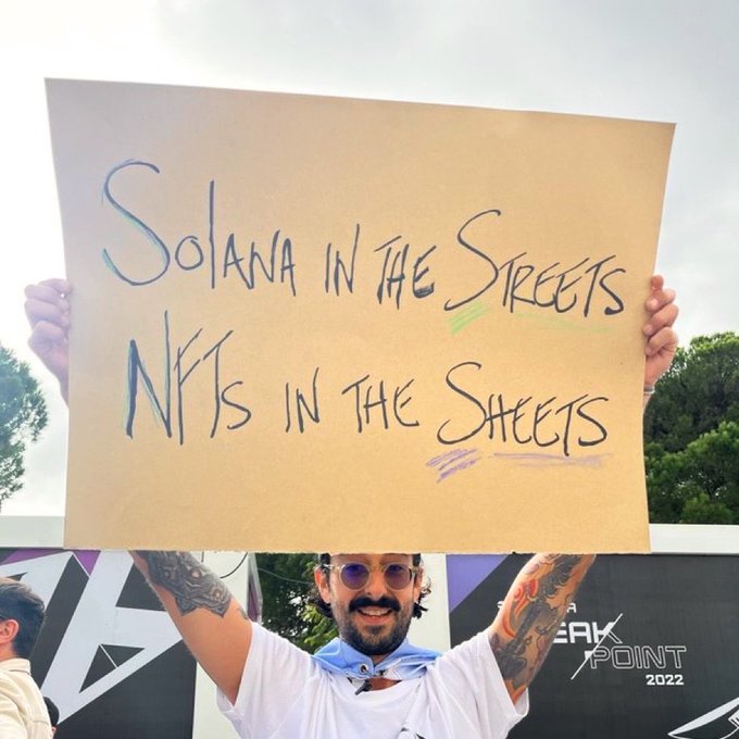 A man with tattoos and sunglasses holds up a cardboard sign reading 'Solana in the Streets NFTs in the Sheets' at what appears to be a Breakpoint 2022 event.