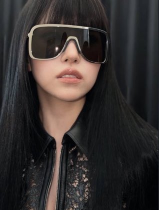 A portrait of a woman with long straight black hair wearing oversized designer sunglasses and a black lace top, photographed against a dark curtain background.