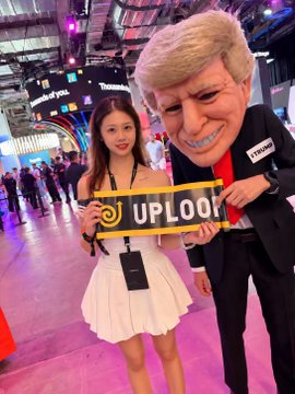 A young woman in a white dress poses at an event beside a person wearing a large caricature head of Donald Trump holding a sign that reads 'UPLook'.