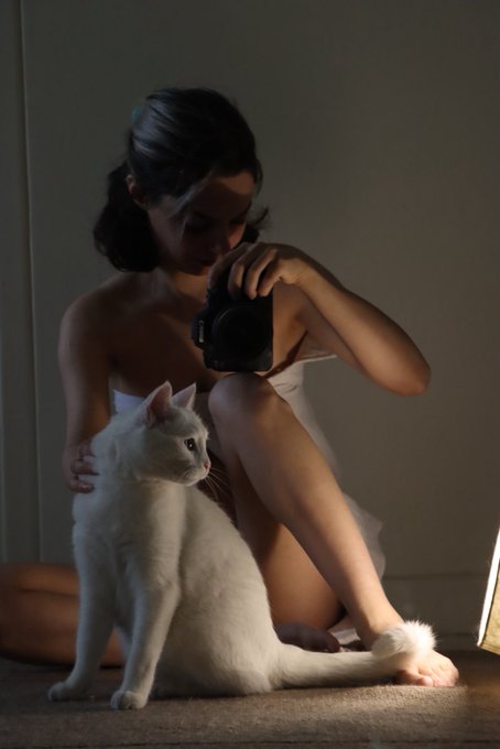 A young woman sits on the floor in a dimly lit room, holding a camera up to her face while a white cat sits in front of her looking toward the viewer.