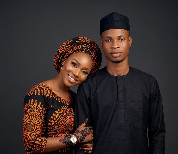 A portrait of a smiling couple in traditional Nigerian attire, with the woman wearing a patterned orange and black headwrap and dress, and the man in a black outfit with a matching cap.
