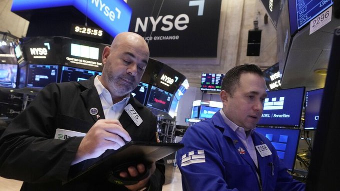 Two NYSE floor traders working amid the busy New York Stock Exchange trading floor, with one trader in a black jacket (badge 119) writing notes and another in a blue jacket (badge 286) monitoring screens.