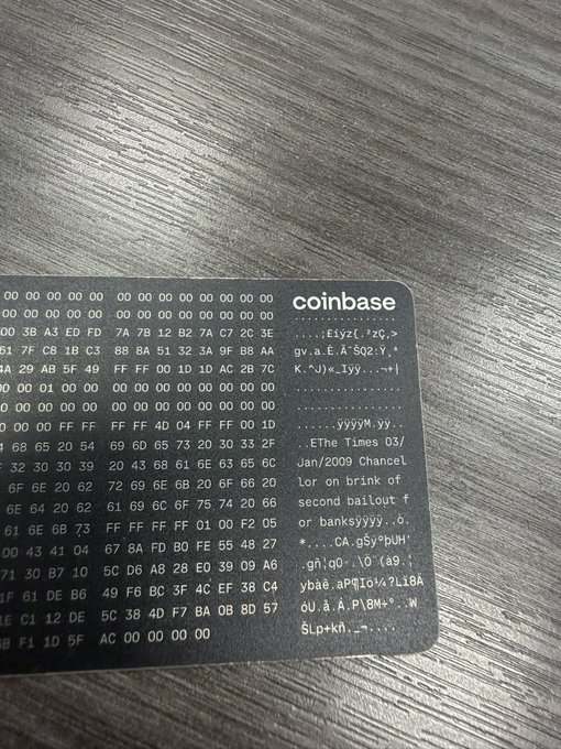 The image shows a coinbase card, which is a small rectangular piece of metal with black text printed on it. The card has a grid-like pattern that displays various numbers and letters. The text on the card reads "Coinbase" followed by a list of numbers and letters in a smaller font size than the main text. The background of the image appears to be a wooden desk or table, providing context for where the coinbase card is being used.