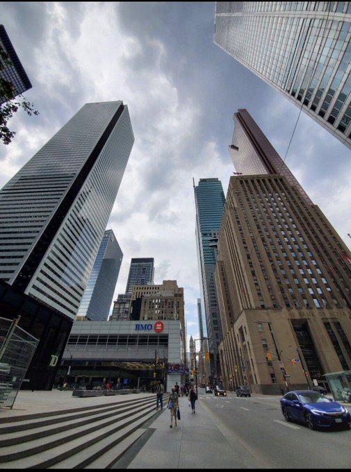 Low-angle photograph of a city street surrounded by tall skyscrapers and office buildings under a cloudy sky, with a BMO bank branch and pedestrians visible at street level.