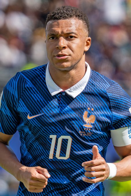 A close-up photograph of soccer player Kylian Mbappé wearing the France national team blue jersey with the number 10 and the FFF crest, with 'PARIS 18.06.2023' visible on the chest.